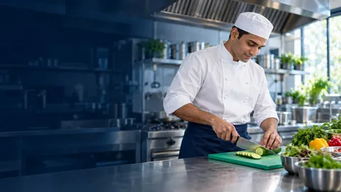 A chef in whites slicing vegetables on a green board in a professional kitchen